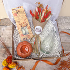 A curated gift box arrangement on a rustic wood surface, featuring a packaged orange candle, a bunch of burnt orange dried flowers labelled Willow Cottage Flowers, a clear glass vase, and an orange spotted glass vase, all nestled in white shredded paper inside a brown box.