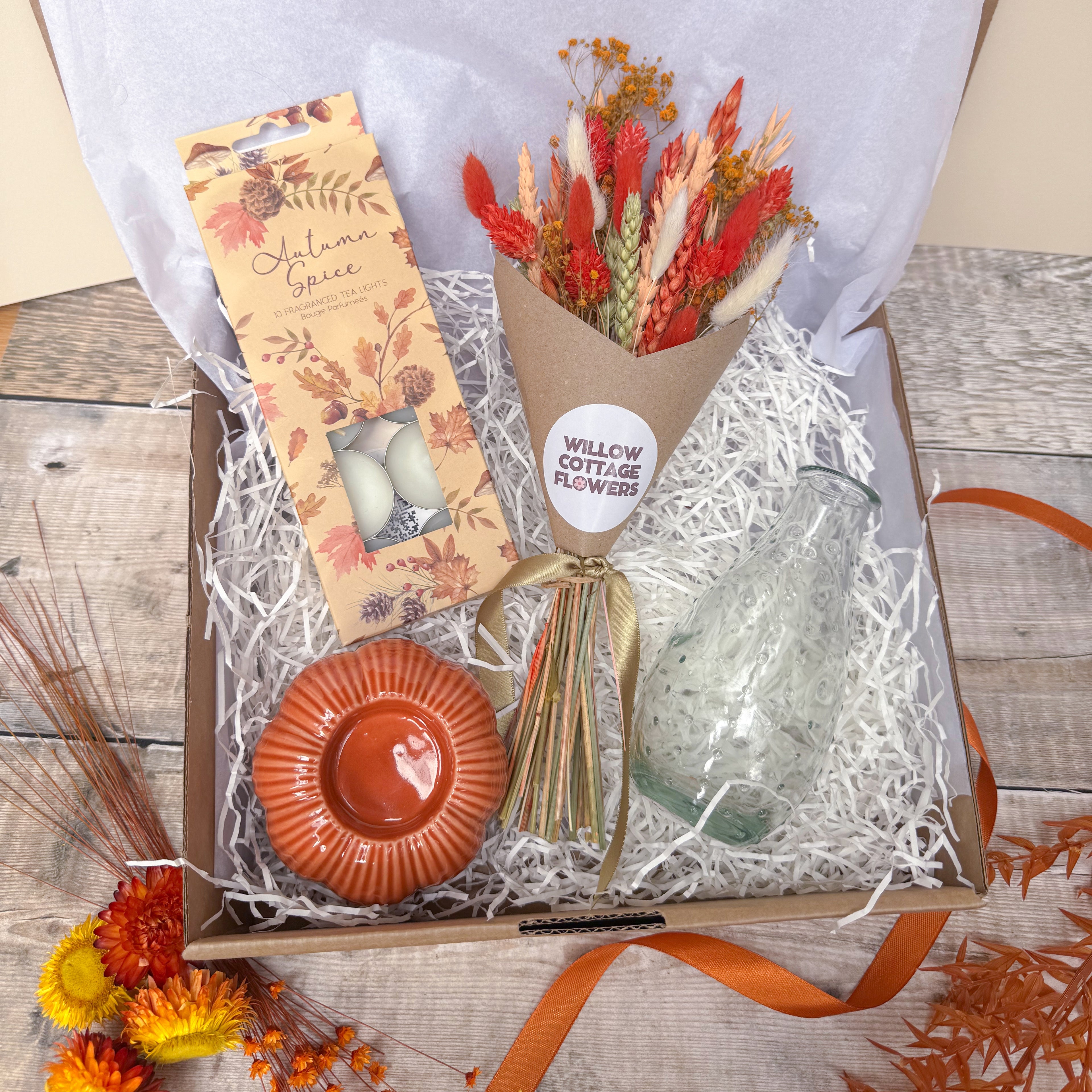 A curated gift box arrangement on a rustic wood surface, featuring a packaged orange candle, a bunch of burnt orange dried flowers labelled Willow Cottage Flowers, a clear glass vase, and an orange spotted glass vase, all nestled in white shredded paper inside a brown box.