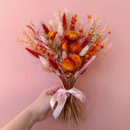 A hand holding a dried flower bouquet with orange, red, pink, and beige botanicals tied with a pale pink ribbon, set against a solid pink background.