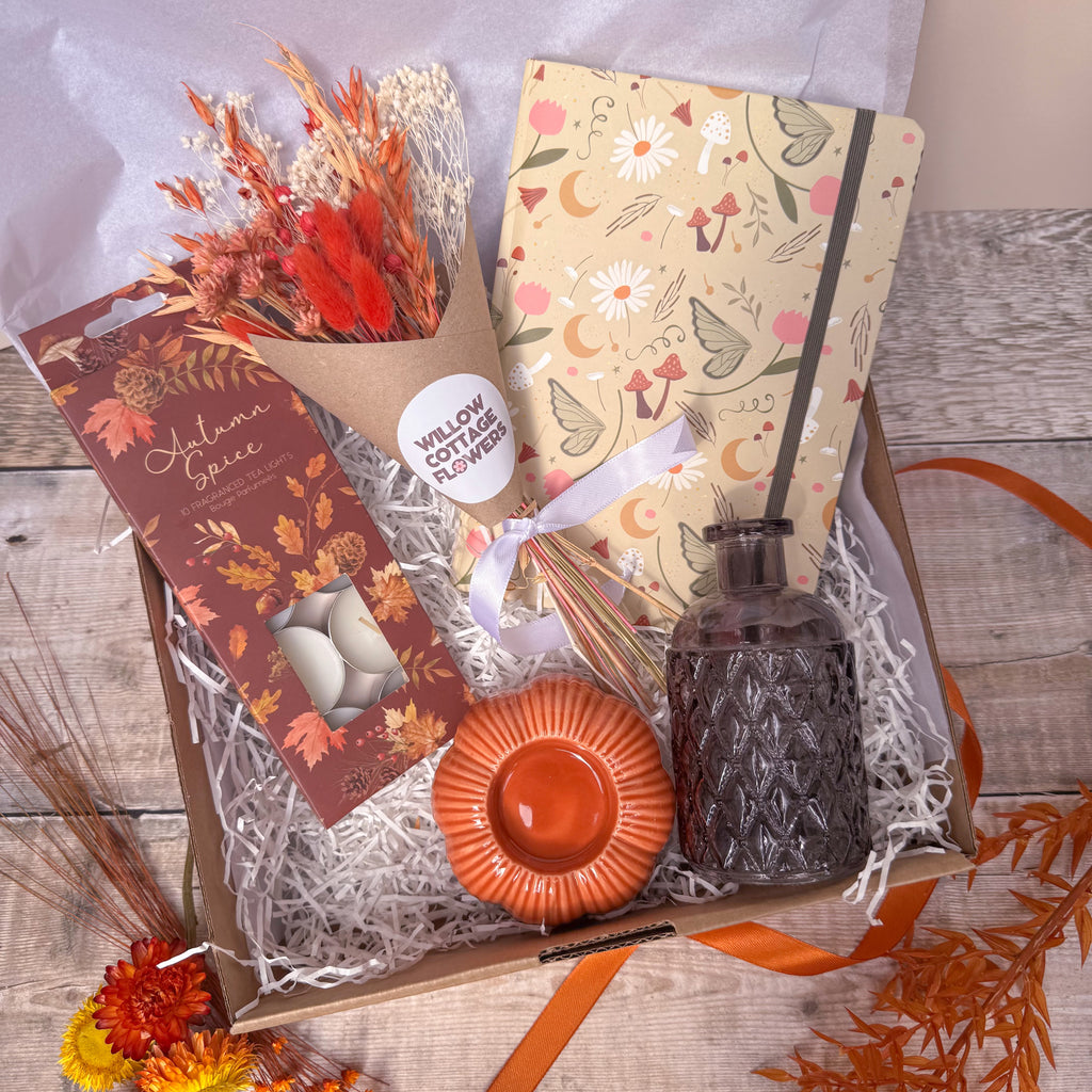 An autumn-themed gift box filled with shredded paper, containing a notebook with floral and fairy patterns, a bunch of dried orange flowers, a box of tealights, a small glass vase and an orange pumpkin shaped candle holder, arranged on a rustic wood surface.