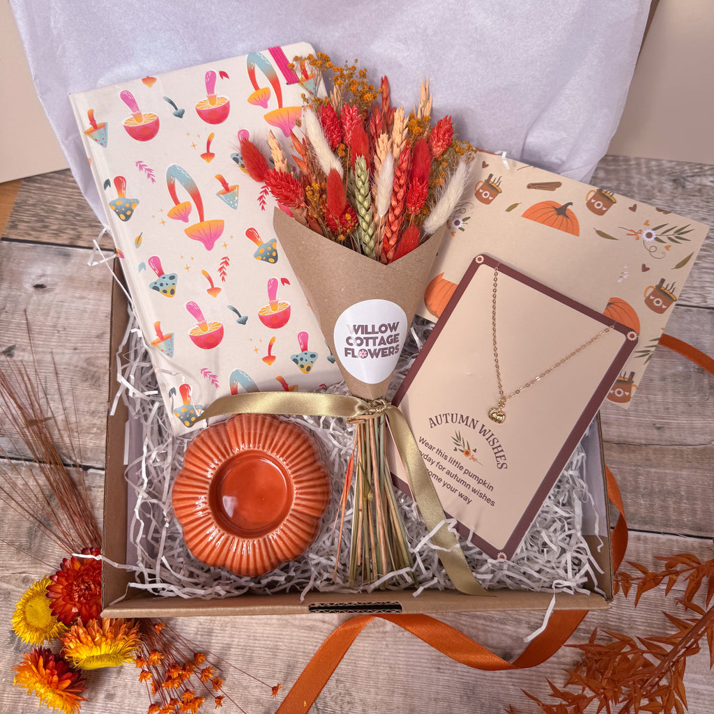 An autumn-themed gift box filled with shredded paper, containing a bouquet of dried wheat and red flowers, a mushroom patterned notebook, a small orange pumpkin tealight holder, a pumpkin charm necklace on a card all arranged on a rustic wooden surface.