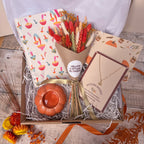 An autumn-themed gift box filled with shredded paper, containing a bouquet of dried wheat and red flowers, a mushroom patterned notebook, a small orange pumpkin tealight holder, a pumpkin charm necklace on a card all arranged on a rustic wooden surface.