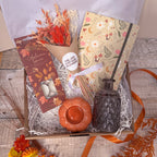 A curated gift box arrangement on a rustic wood surface, featuring a patterned notebook, a dried flower bundle in a cone, an autumn-themed tealight set, a small orange ribbed pumpkin shaped tealight holder, a dark textured glass bottle, and white shredded paper filler inside a gift box.