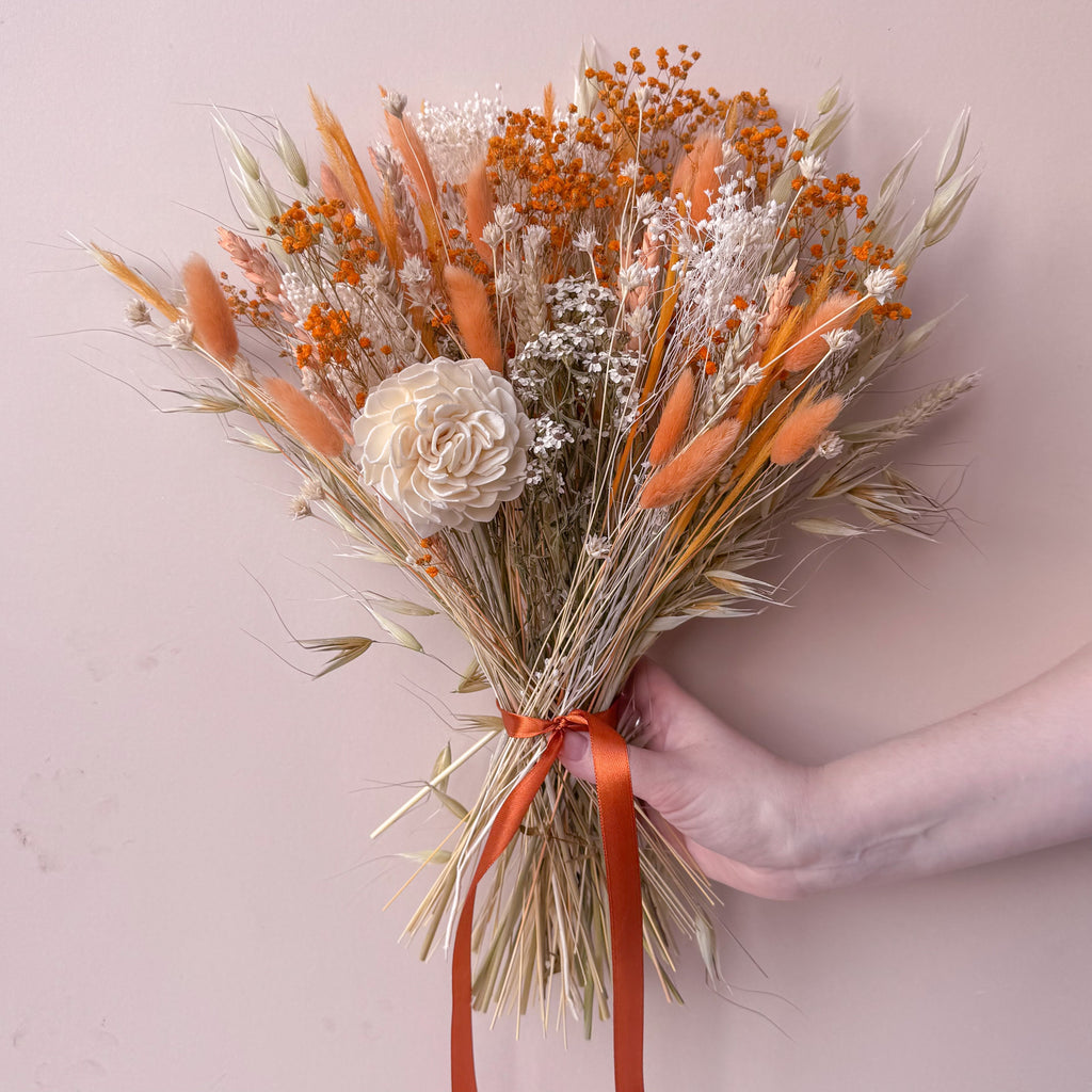 a peach coloured dried flower arrangement with gypsophila, wheat and oats against a plain background