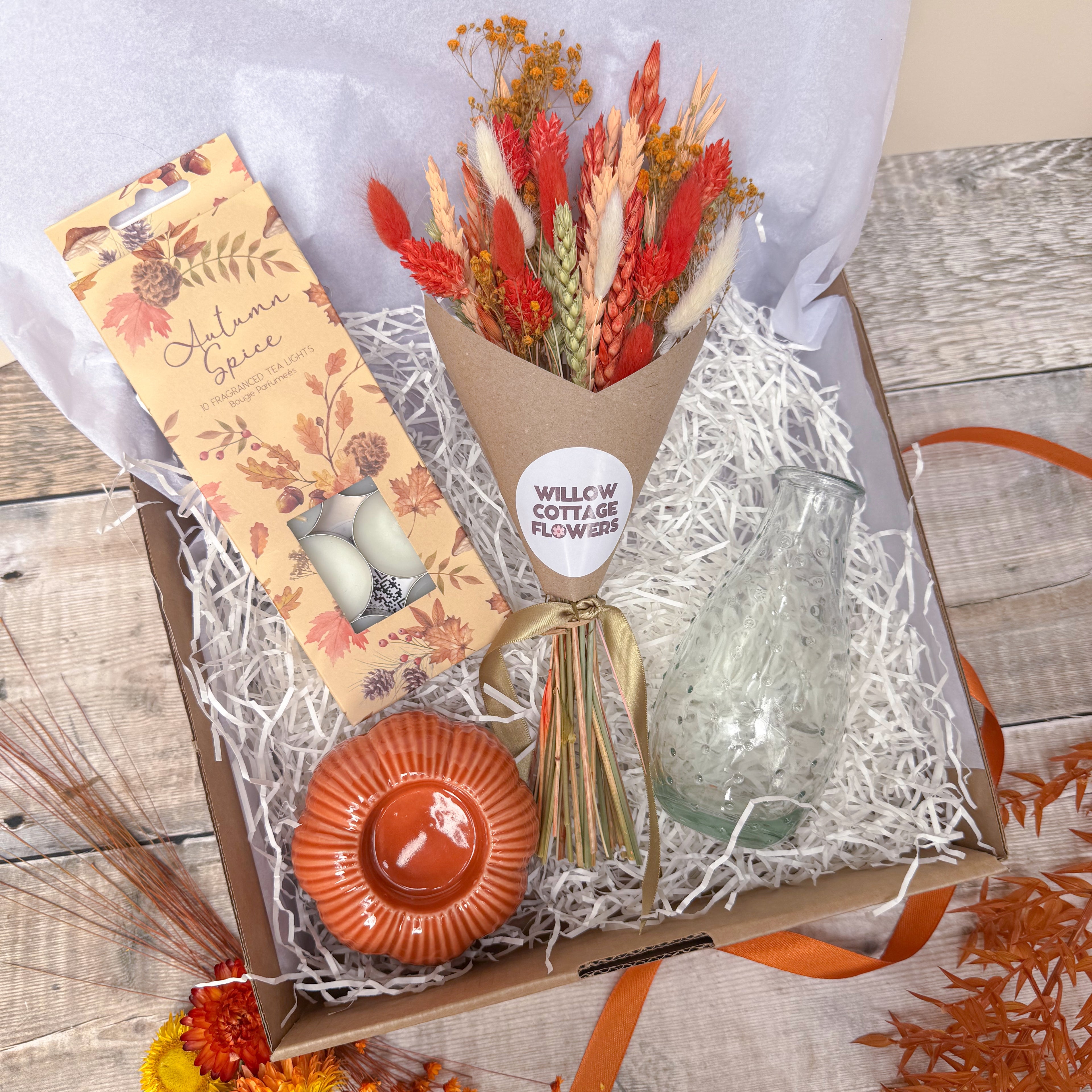 An autumn-themed gift box from Willow Cottage Flowers featuring a dried flower bouquet, autumn spice tea lights, a small orange ceramic pumpkin tea light holder and a clear glass vase, arranged in white paper shred on a rustic wood background.