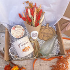 A curated fall gift box filled with seasonal items: a bouquet of dried orange and cream flowers in a brown paper cone, a white enamel mug reading 'Hello Pumpkin,' a round candle tin labeled 'Autumn Spice,' a wooden pumpkin-shaped coaster reading 'Pumpkin Spice,' and a small ribbed glass bottle, all arranged in a cardboard box with shredded paper lining.