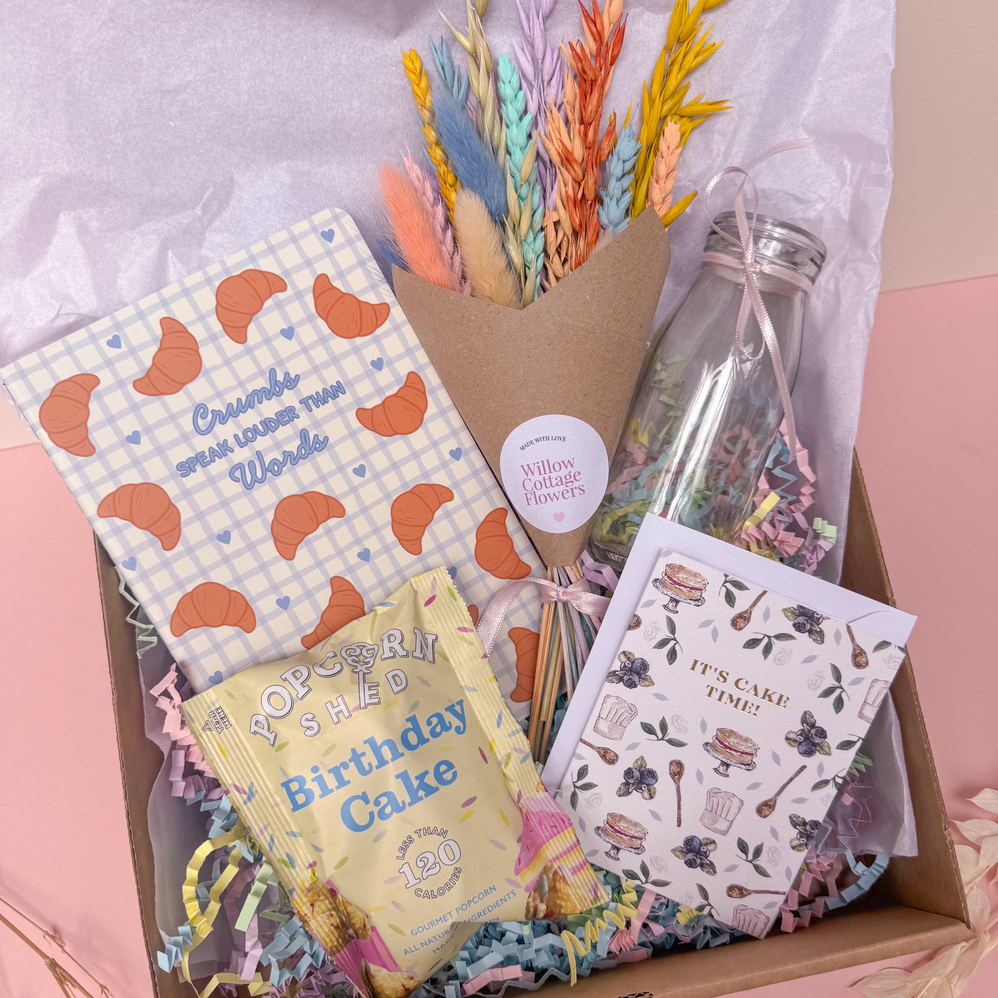 birthday Gift box with stationery items, a bottle of flowers, and a card on a pink surface.