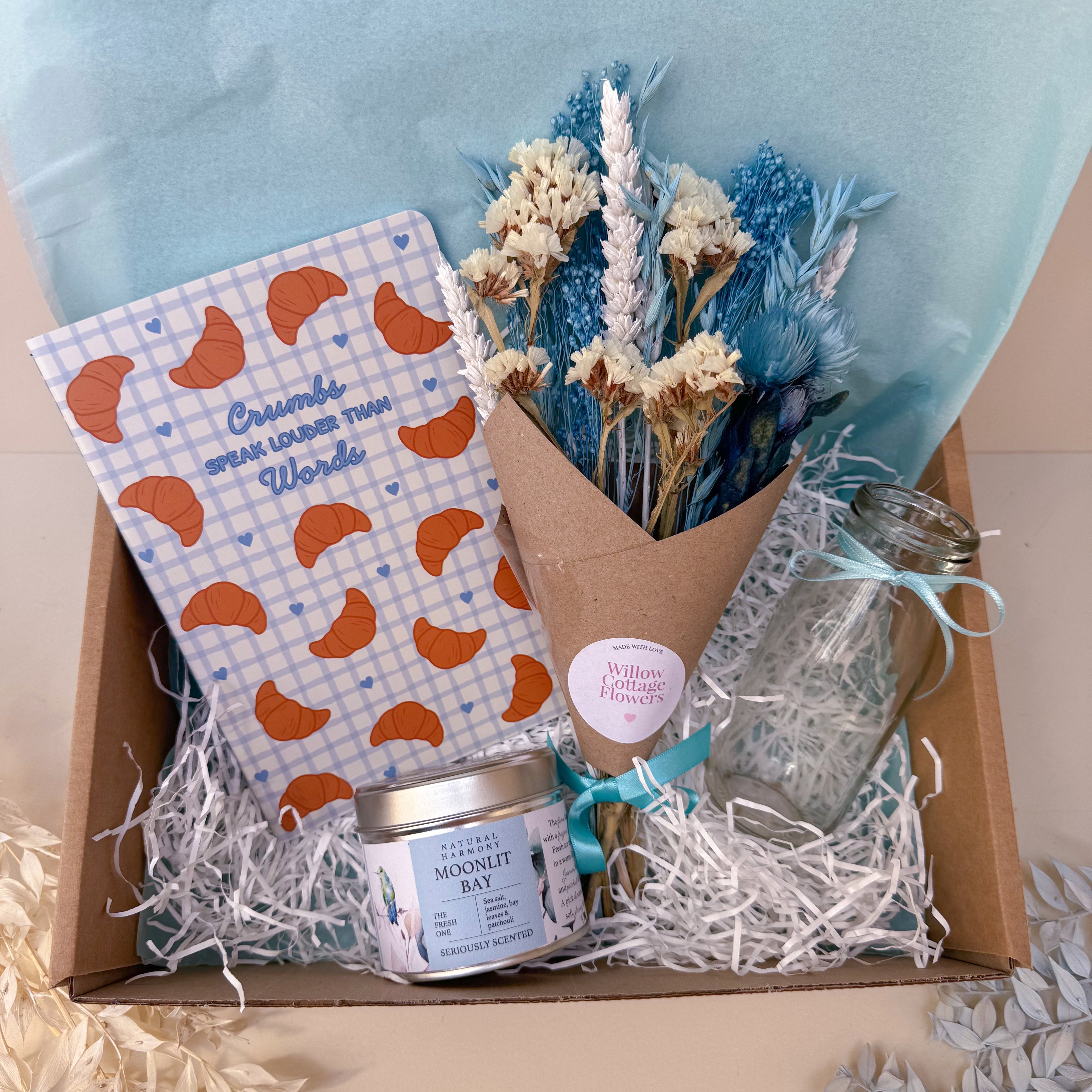 Gift box with a bouquet of flowers, a bottle, a candle, and a croissant notebook inside.