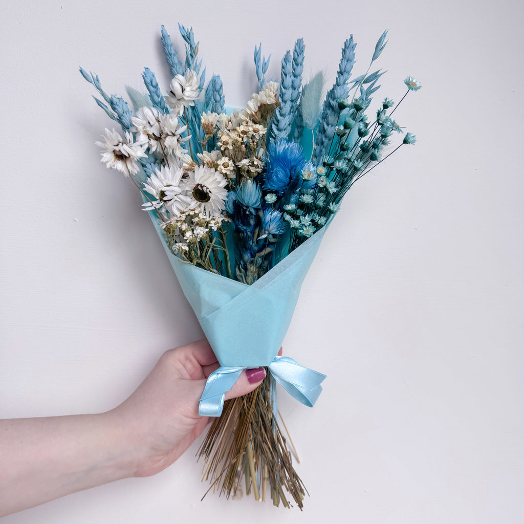 Bouquet of blue and white flowers wrapped in light blue paper held by a hand on a light background