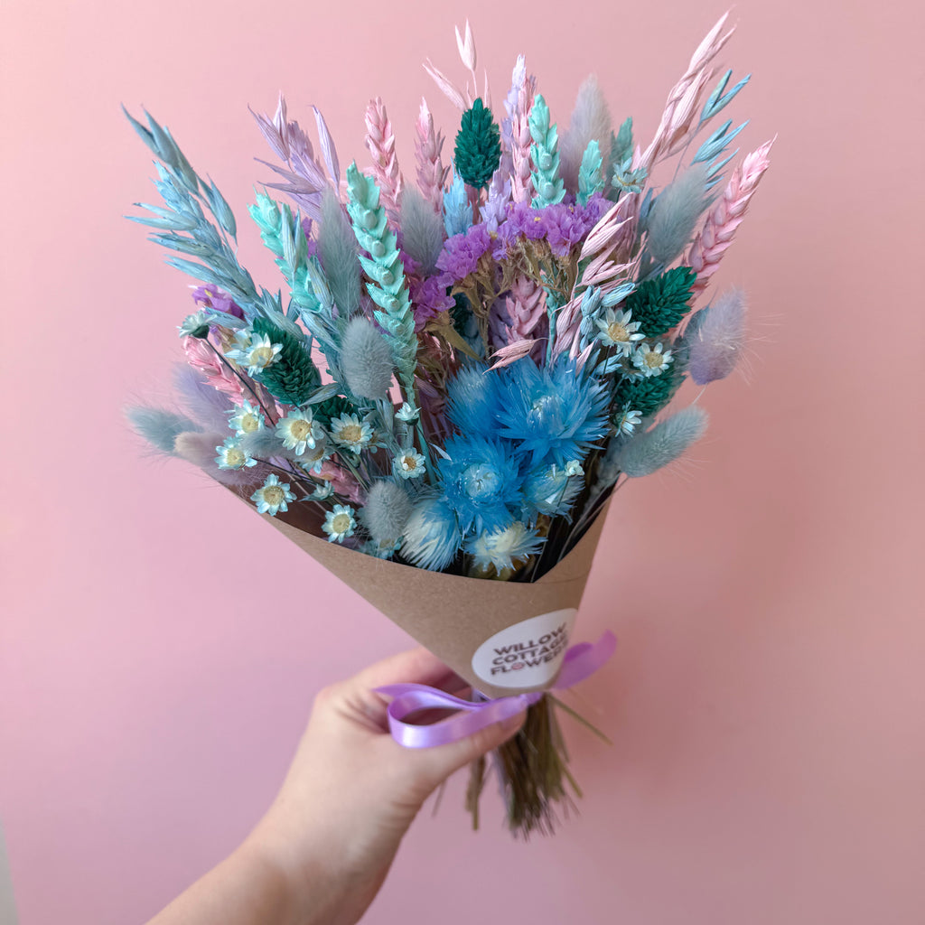 A hand holding a bouquet of dried flowers wrapped in brown kraft paper with a purple ribbon. The bouquet features pastel blue, lilac, aqua, and pink dried grasses and blossoms against a solid pink background.