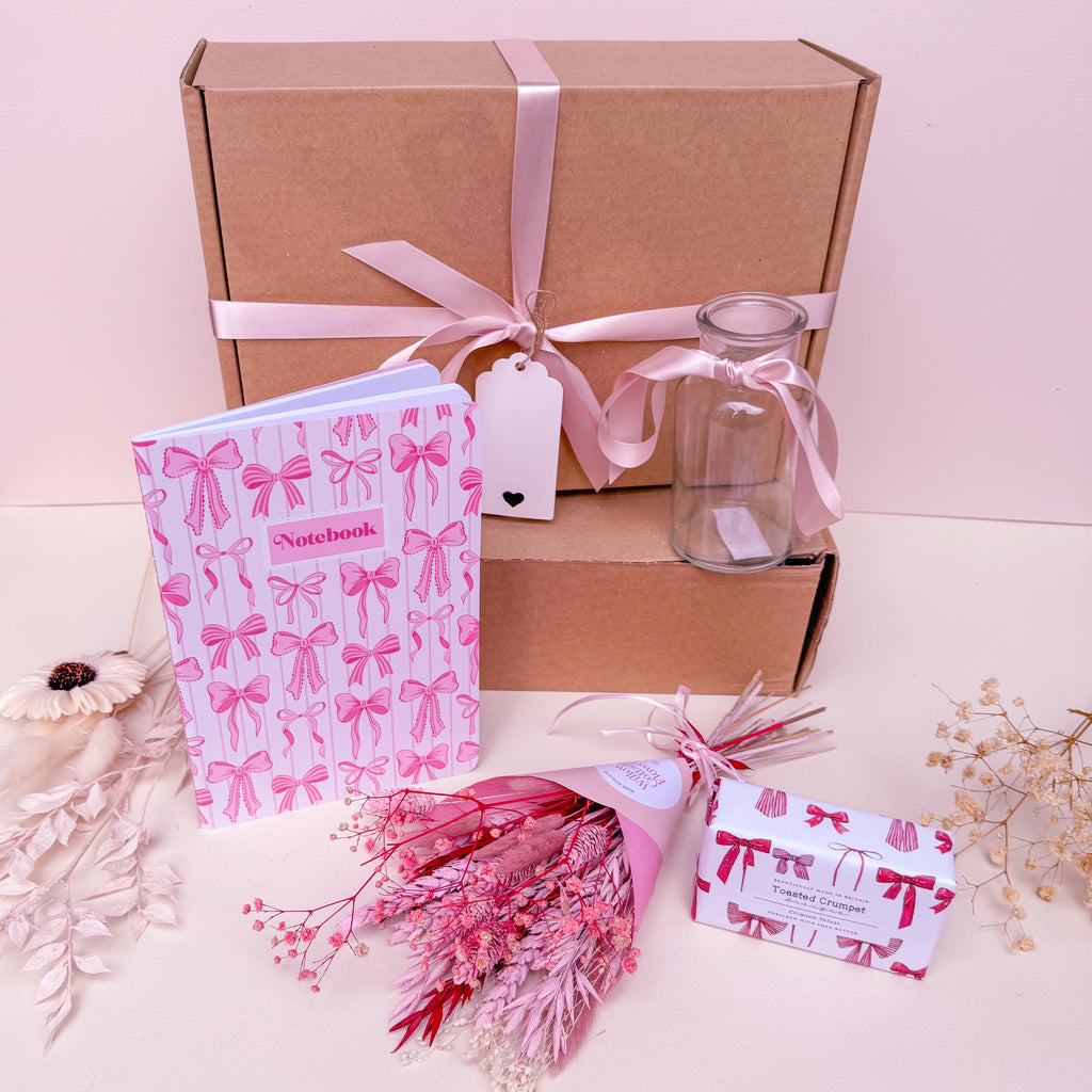 Gift set with a notebook, bouquet, and decorative items on a light pink background in a pink bow theme