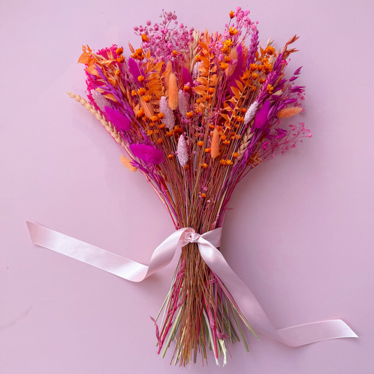 A dried flower bouquet with vibrant orange and pink botanicals tied with a pale pink ribbon, set against a solid light pink background.
