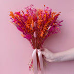 A hand holding a vibrant dried flower bouquet with bright orange, pink, and magenta botanicals tied with a pale pink ribbon, set against a solid pink background.