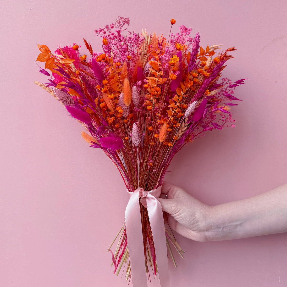 A hand holding a vibrant dried flower bouquet with bright orange, pink, and magenta botanicals tied with a pale pink ribbon, set against a solid pink background.