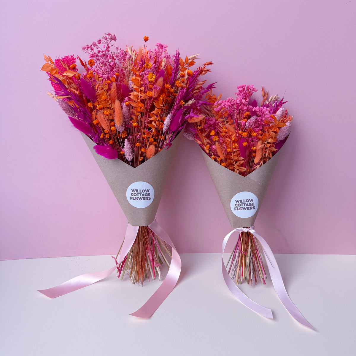 Two brown gift wrapped dried flower bouquets with pink and orange blooms, tied with pale pink ribbons, displayed on a white surface against a pink background. Labelled 'willow cottage flowers'