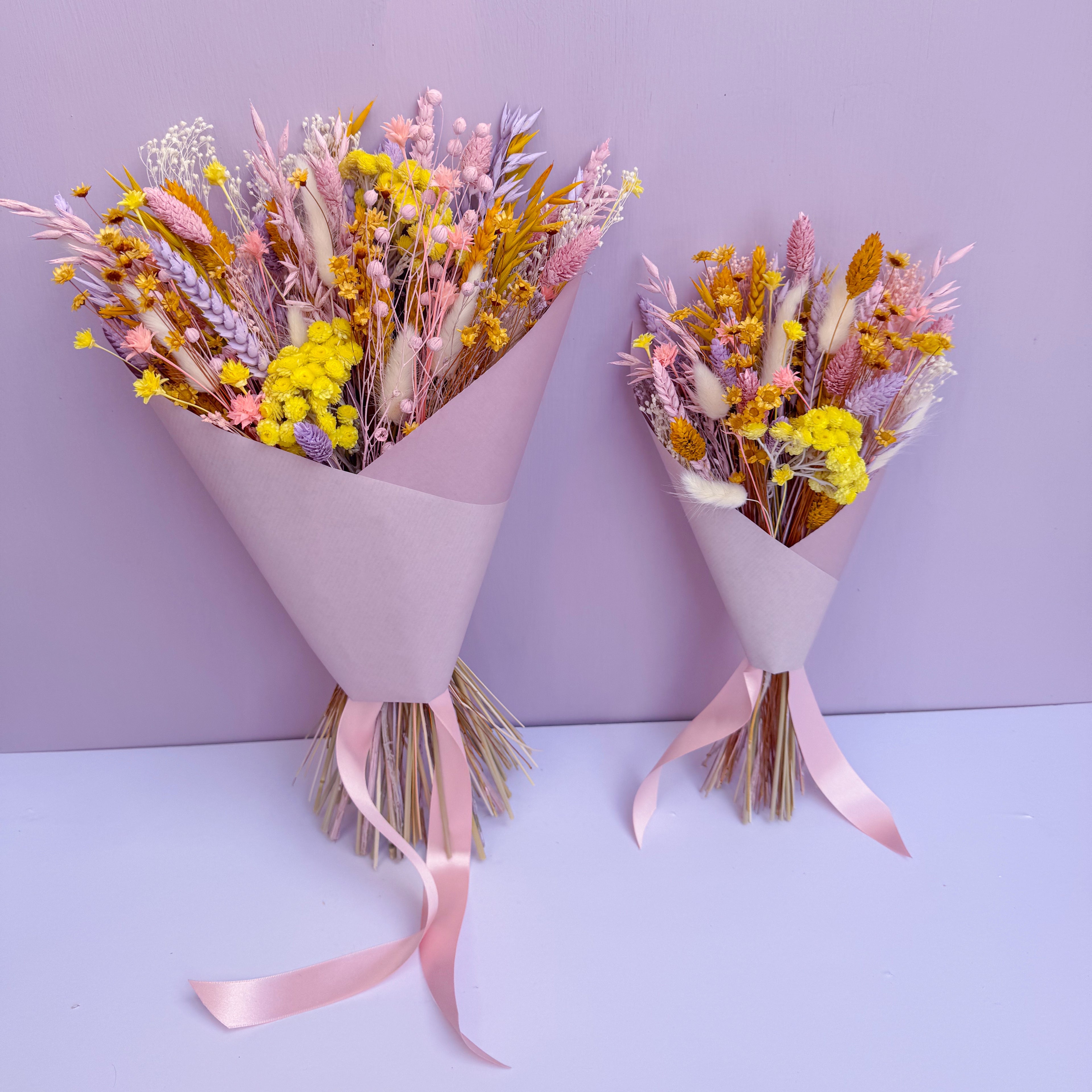 Two bouquets of pastel dried flowers with pink ribbons on a light purple background