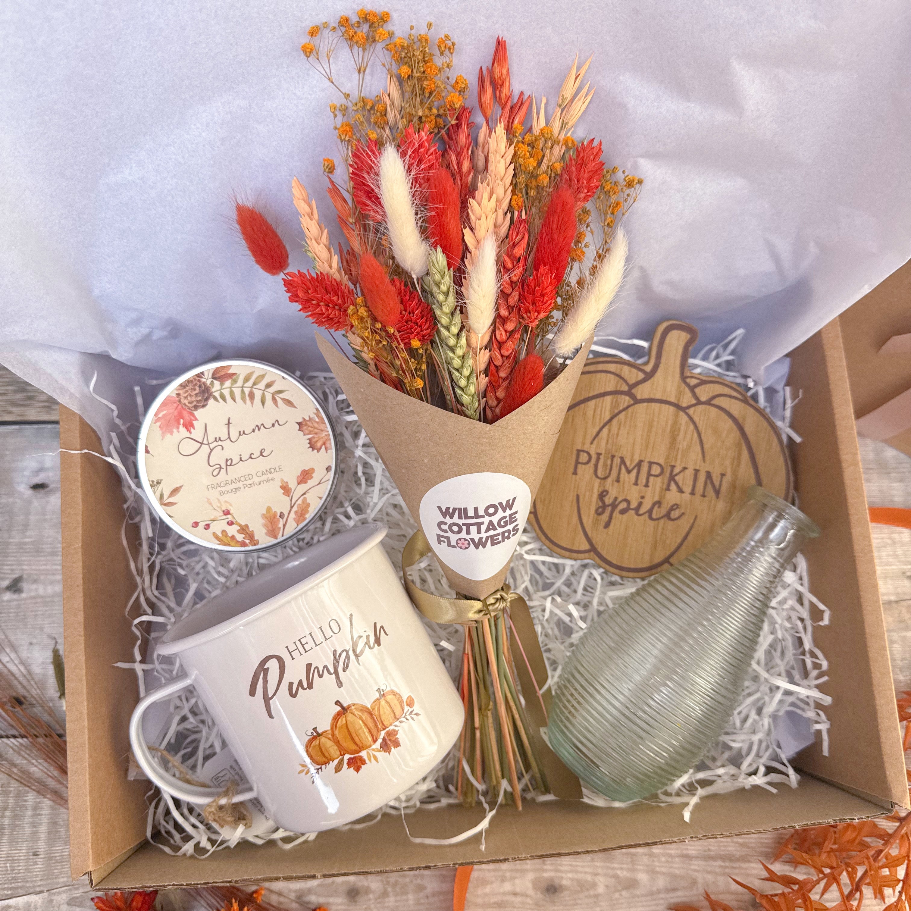 An autumn-themed gift box filled with seasonal items, including a bouquet of dried red, orange, and cream flowers in a paper gift wrap, a white ceramic mug that says 'HELLO Pumpkin', a round candle tin labeled 'Autumn Spice', and a small ribbed glass bottle, all arranged in white paper shred inside a cardboard box.