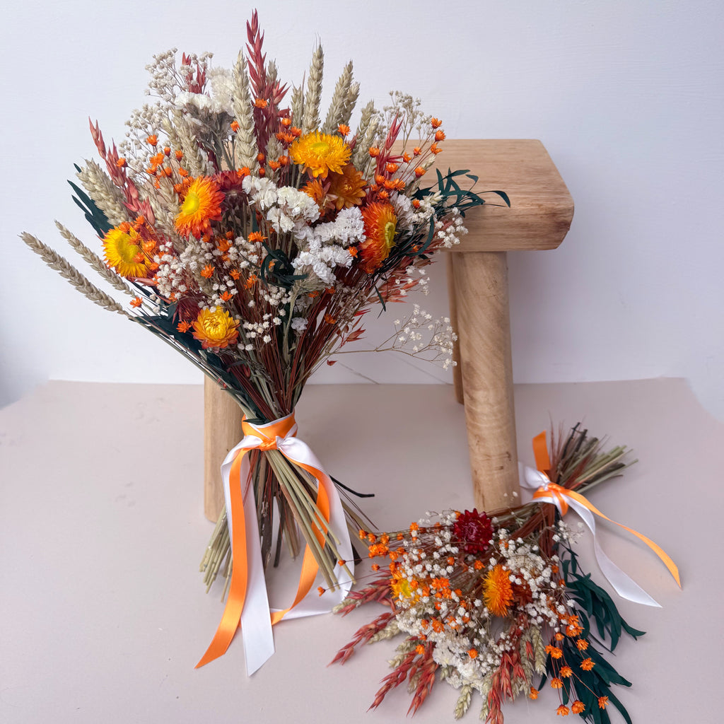 A dried flower bouquet arrangement displayed on a light wood stool and on the floor, featuring orange and yellow wildflowers, white baby's breath, and green stems with orange-and-white ribbons.