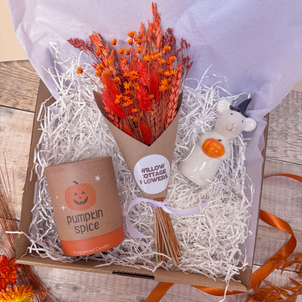 An autumn themed gift box set containing a dried flower bouquet with orange tones, a small ceramic white mouse figurine holding an orange pumpkin, and candle labelled 'pumpkin spice'. The items are arranged in white shredded paper inside a brown box with light purple tissue paper.