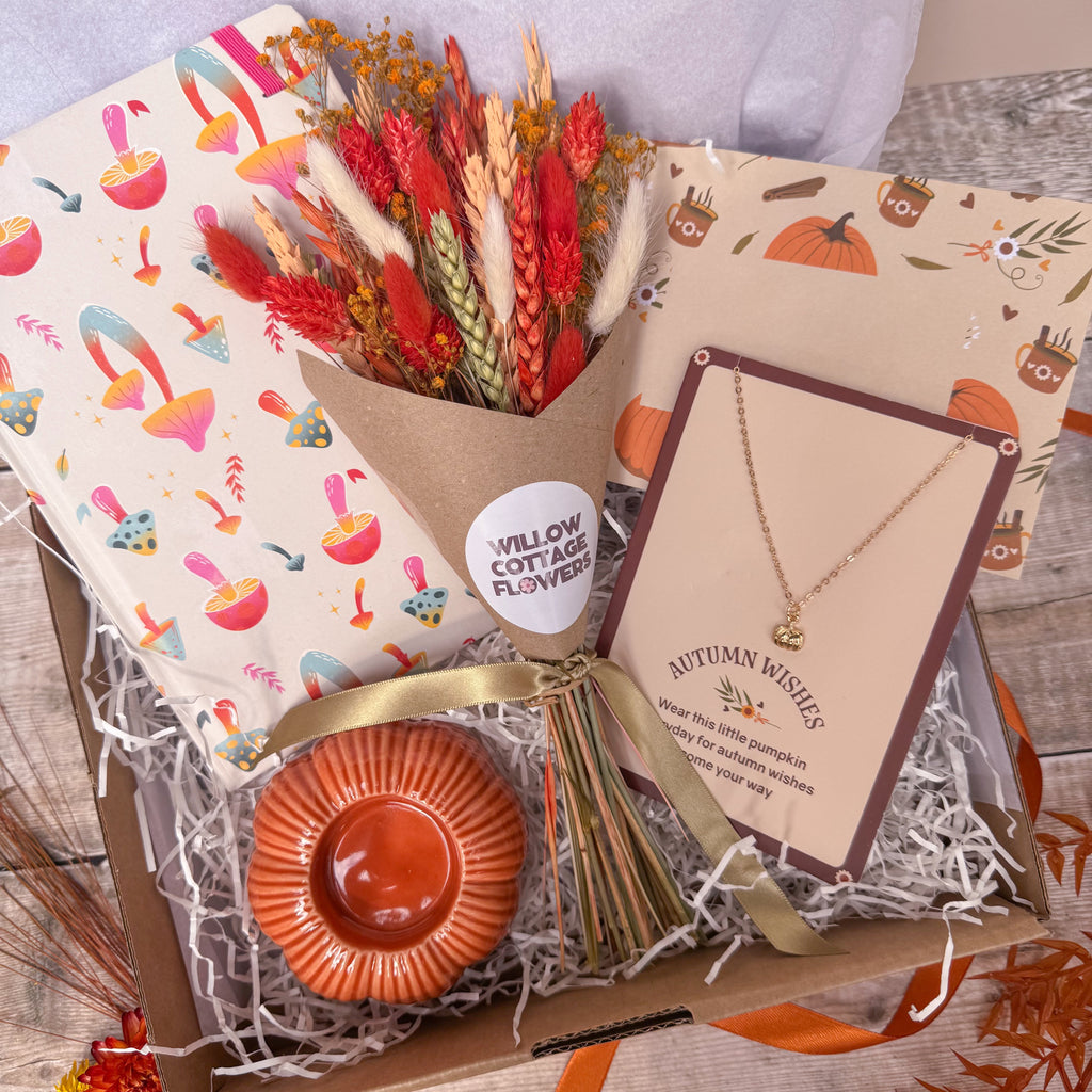 Autumn-themed gift box arrangement featuring a dried flower bouquet in a kraft paper cone, a gold pumpkin charm necklace on a display card, a small orange pumpkin shaped candle holder and mushroom patterned notebook inside a cardboard box with shredded paper filler.