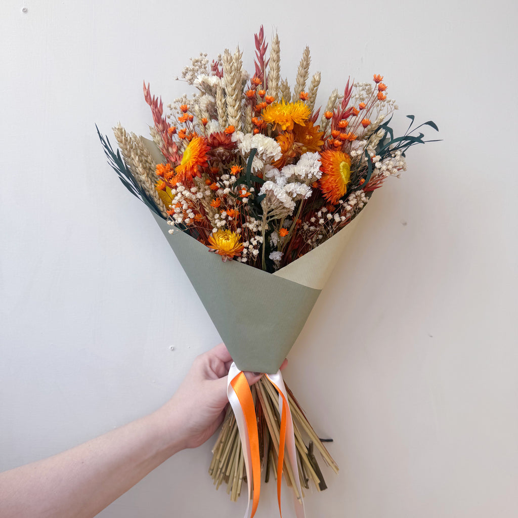 A dried flower bouquet wrapped in a sage green paper sleeve, held by a hand against a plain off-white wall. The bouquet features orange, yellow, and beige dried flowers with green foliage, tied with an orange and white ribbon.