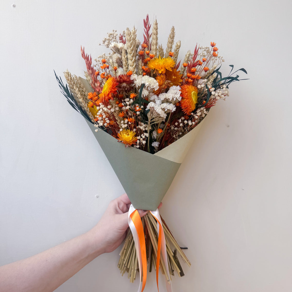 A dried flower bouquet wrapped in a sage green paper sleeve, held by a hand against a plain off-white wall. The bouquet features orange, yellow, and beige dried flowers with green foliage, tied with an orange and white ribbon.