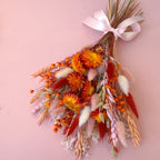 A dried flower bouquet featuring orange strawflowers, oats, and fluffy white bunny tail grass, tied with a pale pink ribbon on a solid pink background.