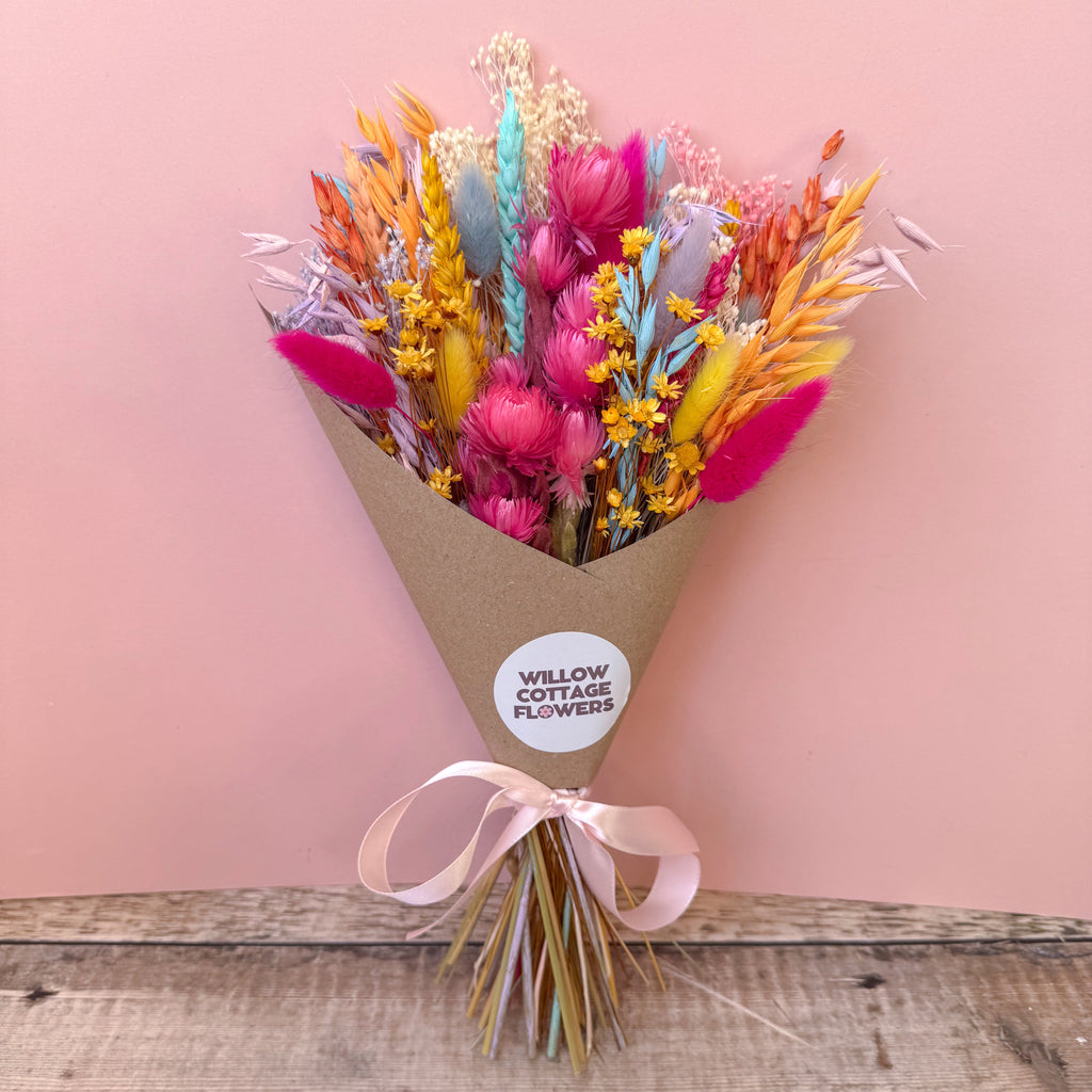 A dried flower bouquet wrapped in brown kraft paper with a pale pink ribbon, featuring bright pink, orange, yellow, blue, and purple flowers, set against a solid pink background on a rustic wooden surface. A round white sticker on the wrapper reads 'WILLOW COTTAGE FLOWERS'.