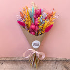 A dried flower bouquet wrapped in brown kraft paper with a pale pink ribbon, featuring bright pink, orange, yellow, blue, and purple flowers, set against a solid pink background on a rustic wooden surface. A round white sticker on the wrapper reads 'WILLOW COTTAGE FLOWERS'.