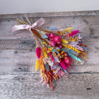A small dried flower bouquet with a pale pink ribbon, featuring bright pink, yellow, orange, blue, and purple botanicals, placed on a weathered gray wooden surface.