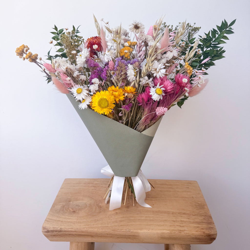 A dried flower bouquet in a sage green cone-shaped wrapper with a white ribbon, displayed on a wooden stool against a plain light background.