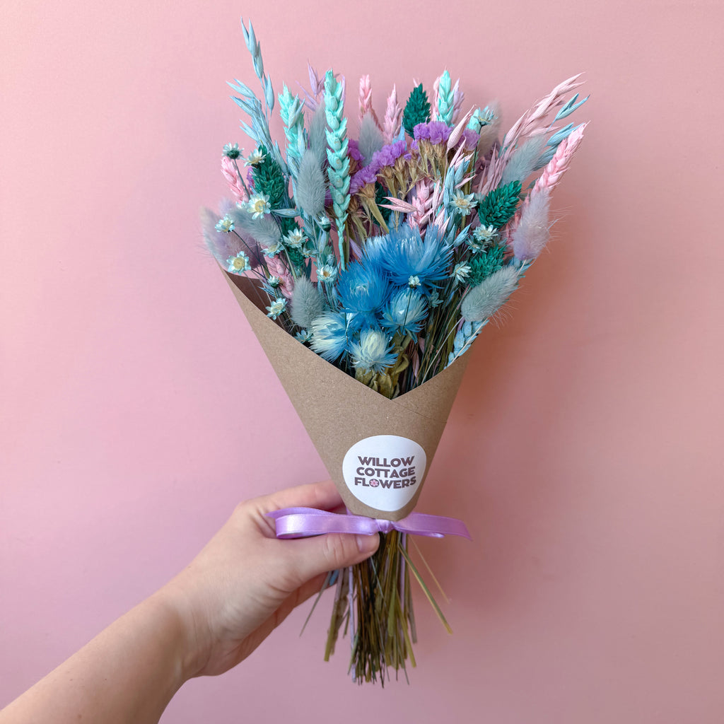 A hand holding a dried flower bouquet wrapped in brown kraft paper with a purple ribbon and a round white sticker that reads 'WILLOW COTTAGE FLOWERS'. The bouquet features pastel blue, pink, and green dried flowers against a solid pink background.