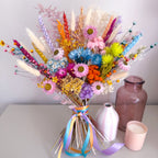 A vibrant dried flower bouquet in a clear glass vase with a rainbow ribbon, styled on a light gray surface next to a pink candle, a white vase, and a brown glass bottle.