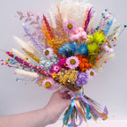 A hand holding a vibrant, multicolor dried flower bouquet with pastel and bright blooms, fluffy grasses, and ribbon ties against a plain light gray background.