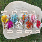 Six small dried flower buttonholes arranged on a round natural wood slice outdoors on grass. Each bouquet is labeled with a color name: Yellow, Blue, Rainbow, Pink, White, Orange, and Red.