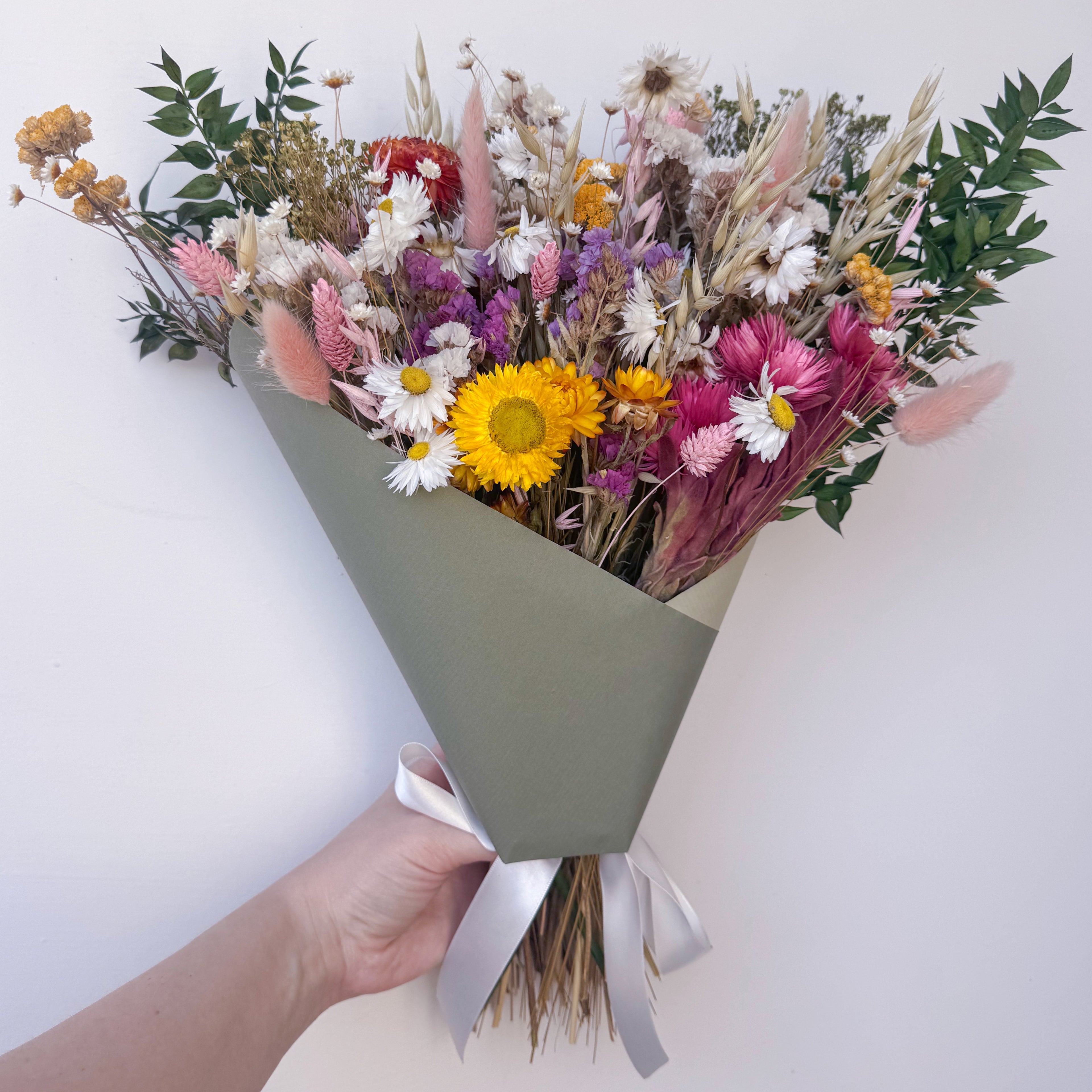 A hand holding a dried flower bouquet wrapped in sage green paper with a white ribbon, featuring pink bunny tails, white and yellow daisies, and green foliage against a plain white background.