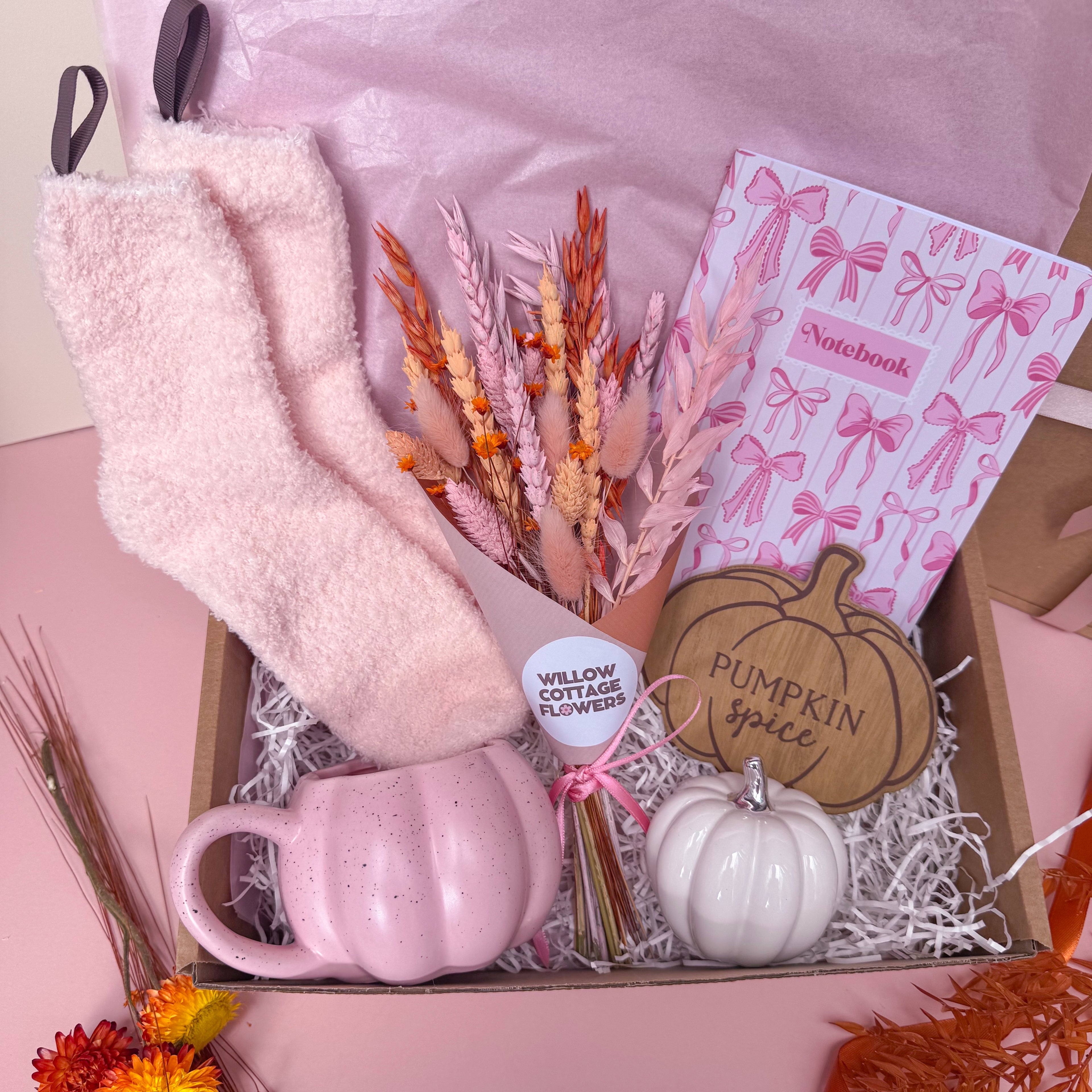 A cosy autumn gift box filled with pink and  autumn themed items, including fluffy pink socks, a pink notebook with bow patterns, a pink speckled pumpkin shaped mug, dried flower bunch, a small white pumpkin ornament, and a wooden pumpkin coaster reading 'Pumpkin Spice' .