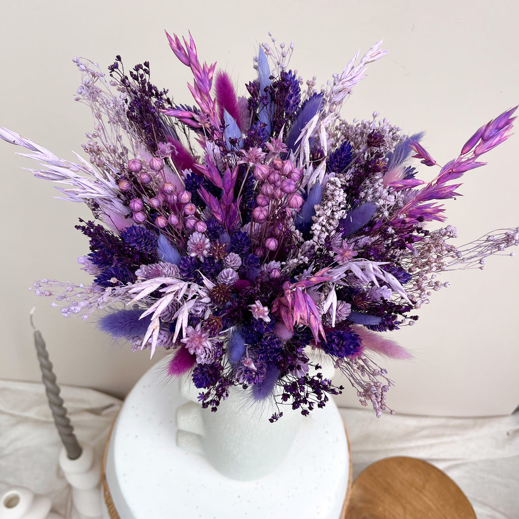 A dried flower arrangement in a white speckled ceramic vase, featuring shades of purple, lavender, and magenta botanicals, set against a neutral beige background.