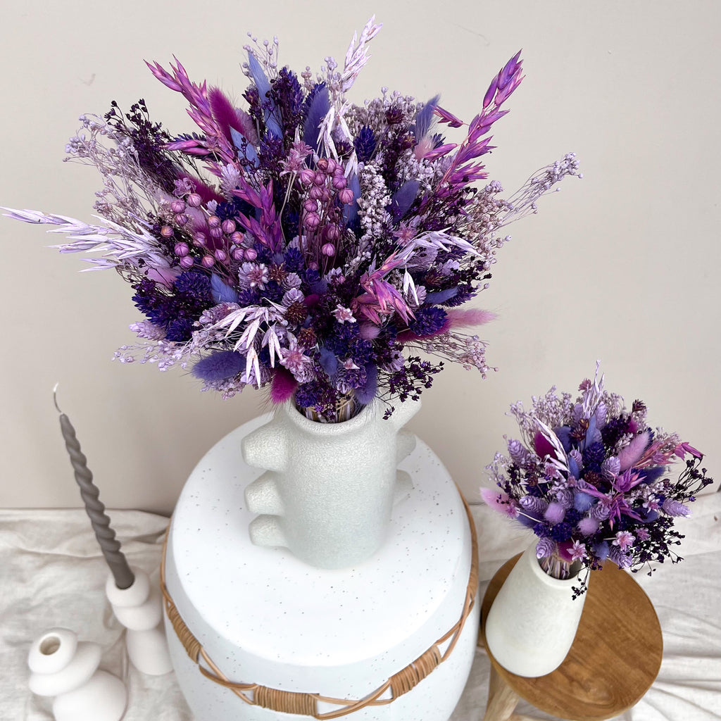 Purple and violet dried flower arrangements in white ceramic vases, styled on a tabletop with a candle and a small wooden stool against a plain beige wall.