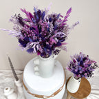 Purple and violet dried flower arrangements in white ceramic vases, styled on a tabletop with a candle and a small wooden stool against a plain beige wall.