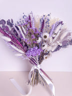 A dried flower bouquet featuring lavender, purple, white, and pink botanicals tied with a white satin ribbon, set against a pale pink background.
