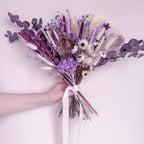 A hand holding a dried flower bouquet with lavender, purple, white, and lilac elements tied with a white ribbon, set against a soft light pink background.