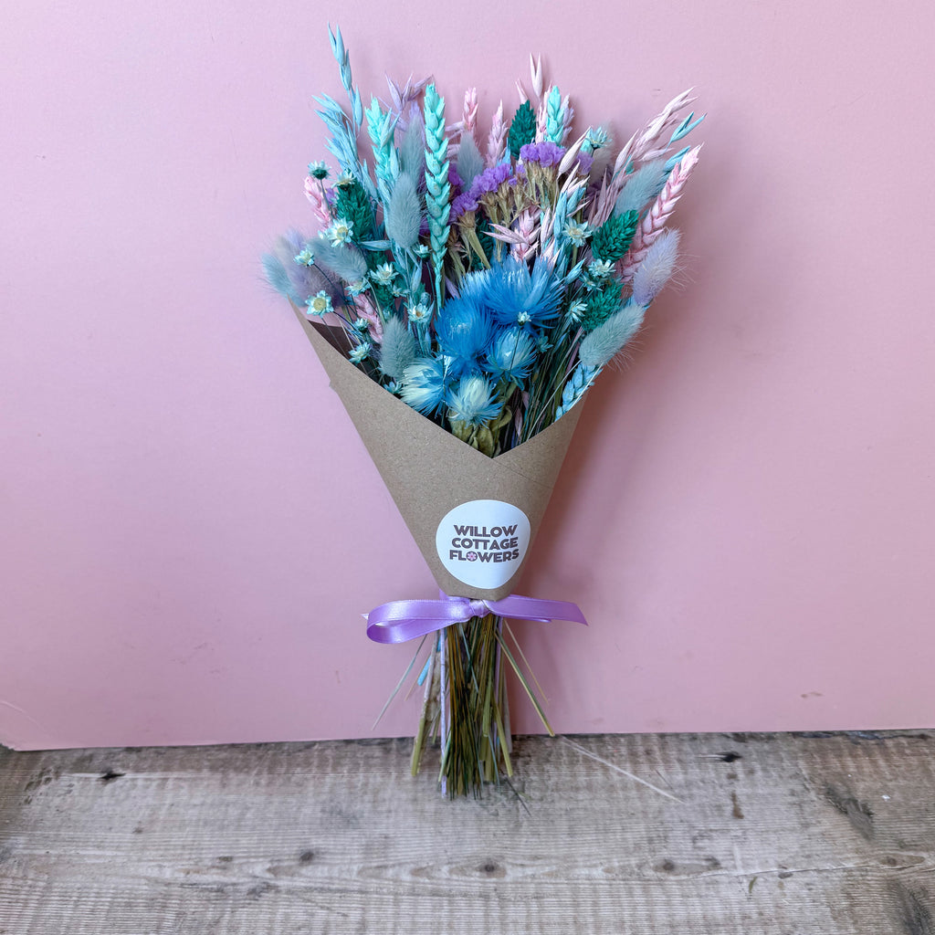 A dried flower bouquet wrapped in brown kraft paper with a purple ribbon and a round white sticker reading 'WILLOW COTTAGE FLOWERS', set against a pink wall and rustic wooden floor.