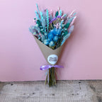 A dried flower bouquet wrapped in brown kraft paper with a purple ribbon and a round white sticker reading 'WILLOW COTTAGE FLOWERS', set against a pink wall and rustic wooden floor.