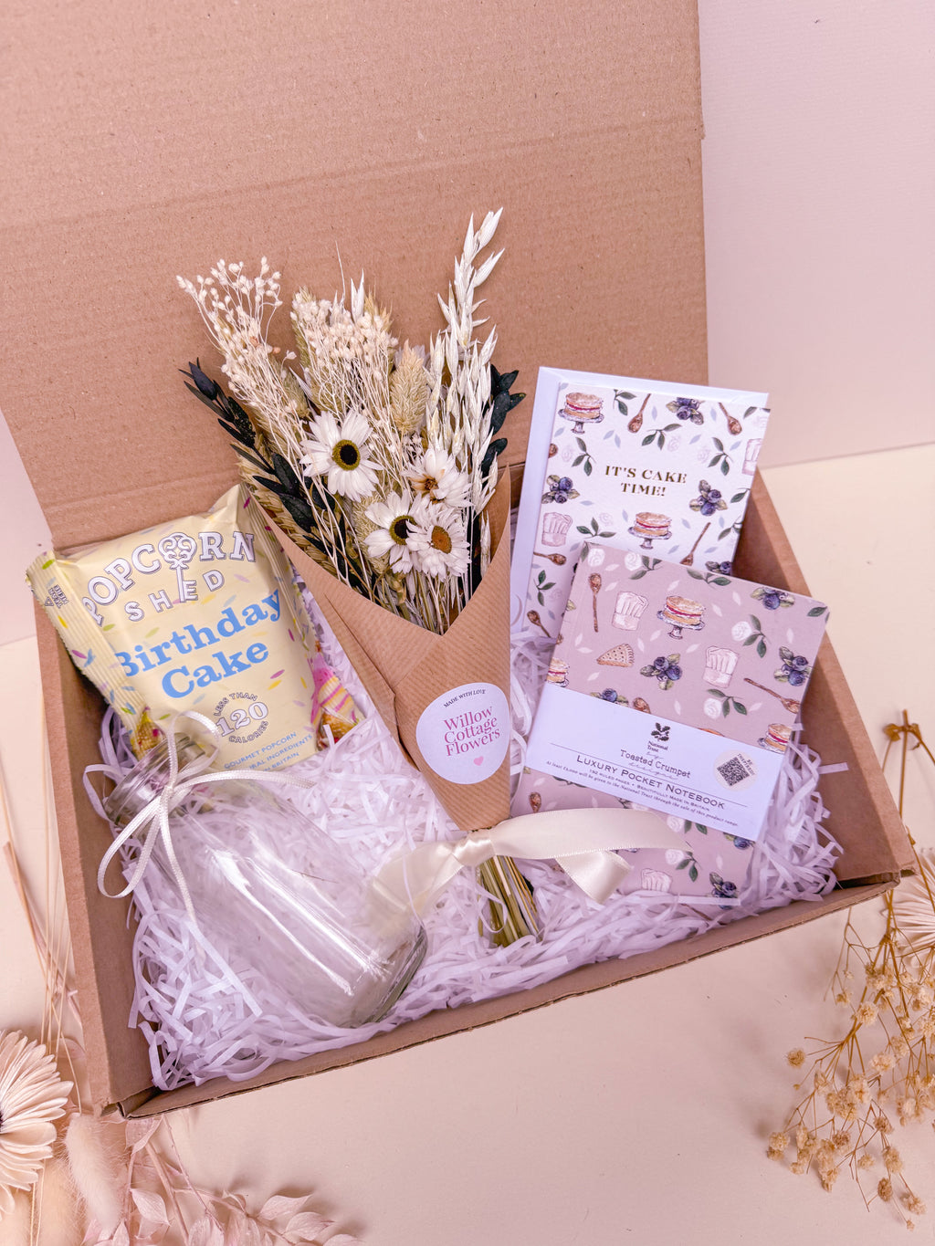Gift box with birthday-themed items including notebook, vase, birthday card, and dried flower bouquet on a light pink background.