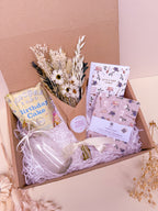 Gift box with birthday-themed items including notebook, vase, birthday card, and dried flower bouquet on a light pink background.