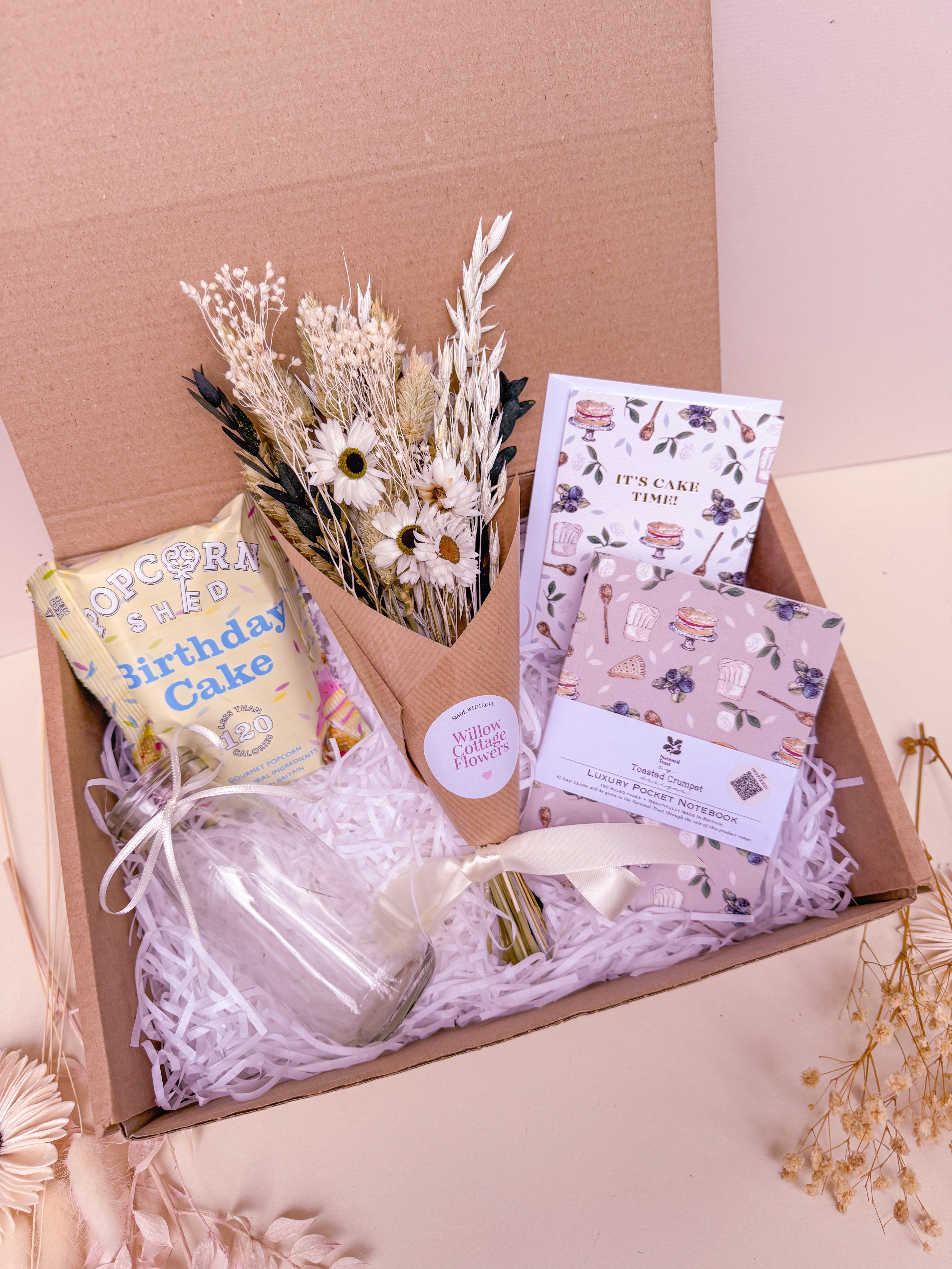 Gift box with birthday-themed items including notebook, vase, birthday card, and dried flower bouquet on a light pink background.