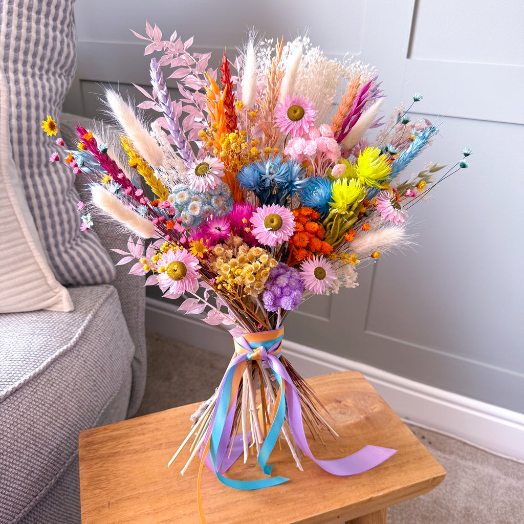 A vibrant, multicolored dried flower bouquet with pink daisies and colourful ribbons, displayed on a wooden stool next to a grey armchair.