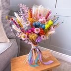 A vibrant, multicolored dried flower bouquet with pink daisies and colourful ribbons, displayed on a wooden stool next to a grey armchair.