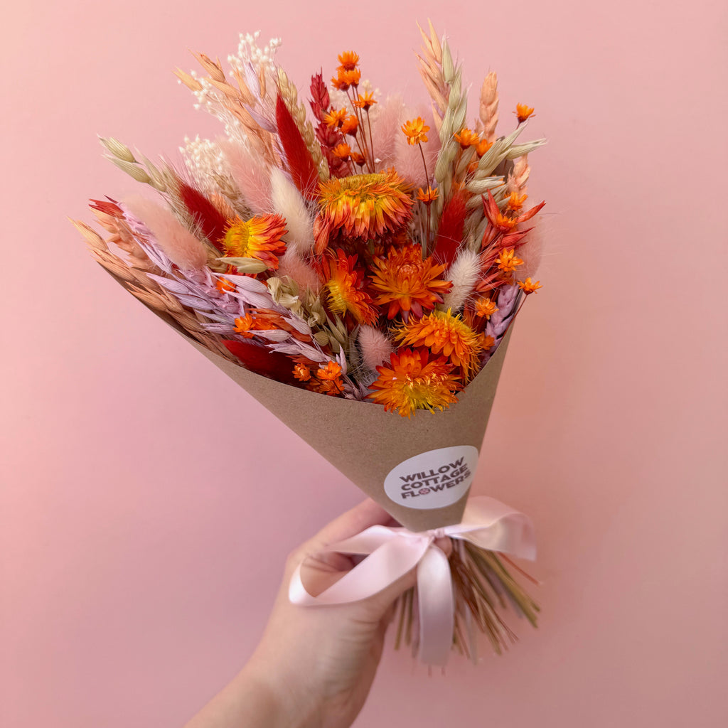A hand holding a dried flower bouquet wrapped in brown kraft paper with a pale pink ribbon. The bouquet features orange, red, beige, and white dried flowers and grasses, with a round white sticker that reads 'WILLOW COTTAGE FLOWERS'.